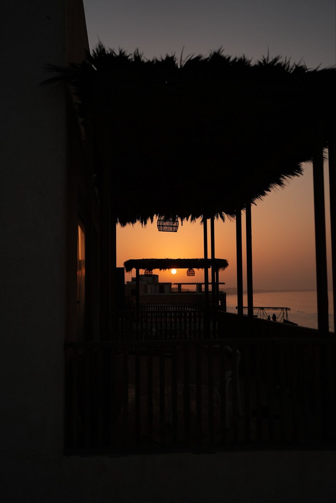 Sun setting over the sea, framed by thatched pergolas and rattan lanterns
